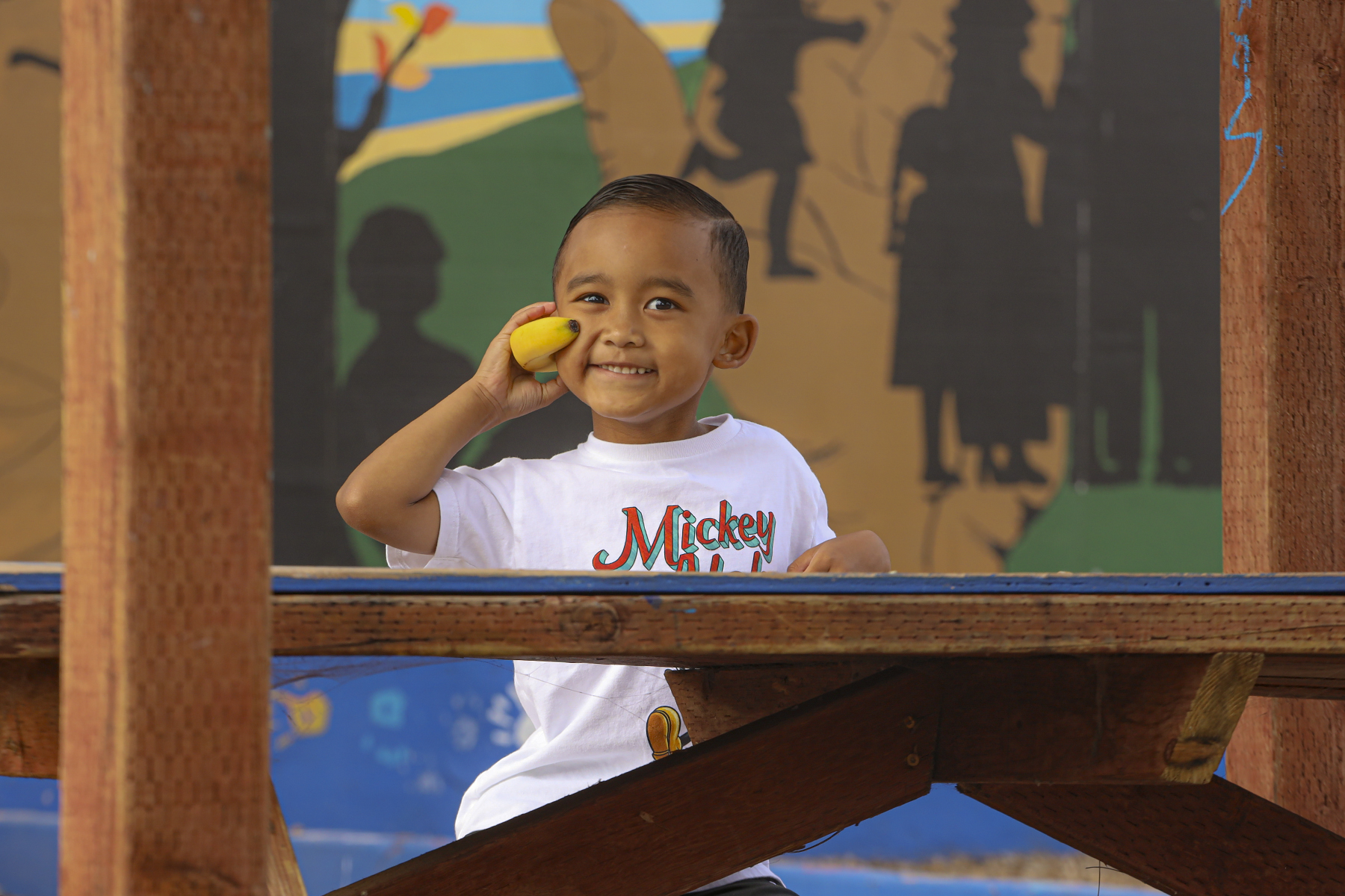 Little boy using a banana as a phone, sitting on a picnic bench in the park